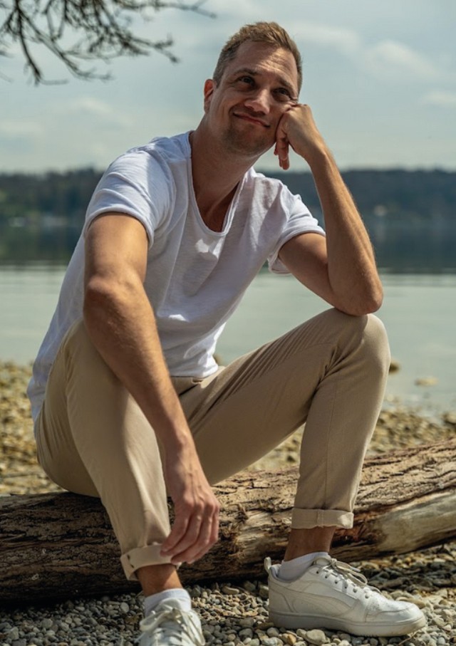 Dominik Hollop sitzt nachdenklich auf einem Baumstamm am Seeufer, trägt ein weißes T-Shirt, beige Hose und weiße Sneaker.