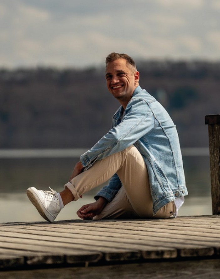 Dominik Hollop sitzt seitlich auf einem Holzsteg am Wasser, trägt eine Jeansjacke und lächelt.