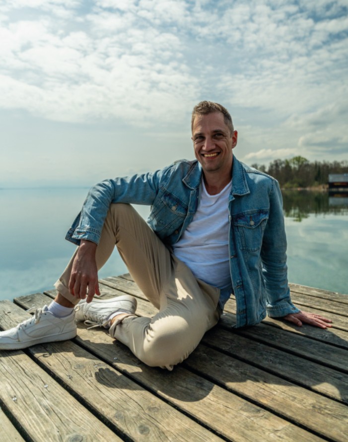 Dominik Hollop sitzt auf einem Steg am Wasser, trägt Jeansjacke und helle Hose, und schaut ruhig in die Natur.