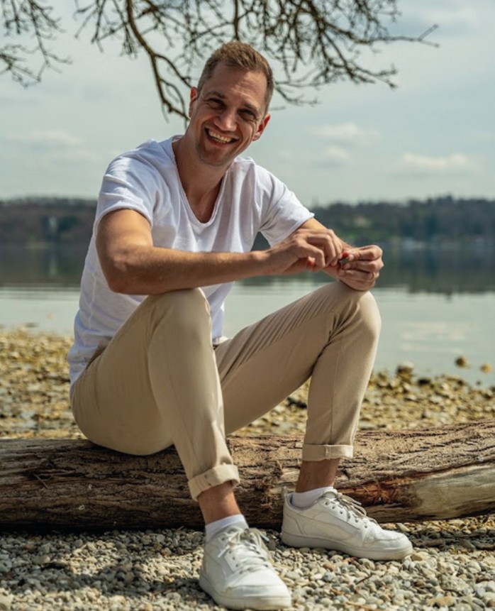 Dominik Hollop sitzt lächelnd auf einem Baumstamm am Seeufer, trägt ein weißes T-Shirt und helle Hosen