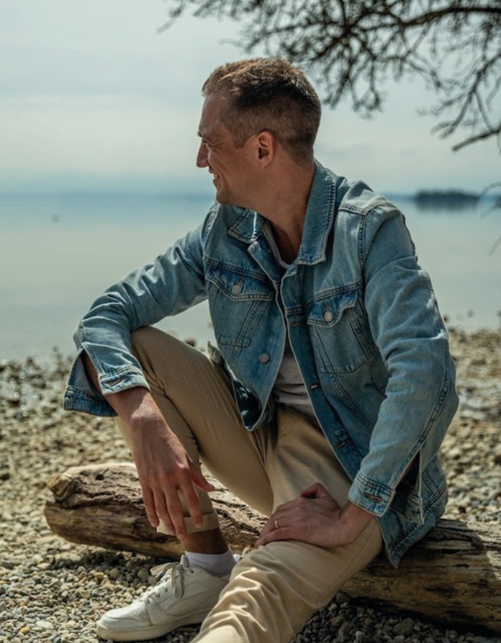 Dominik Hollop sitzt auf einem Baumstamm am Seeufer, trägt Jeansjacke und schaut gedankenvoll in die Ferne.
