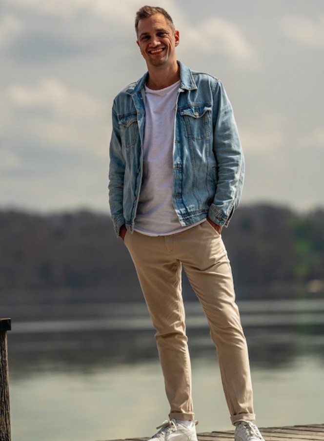 Dominik Hollop steht auf einem Steg am Wasser, trägt Jeansjacke und helle Hose, und lächelt entspannt.