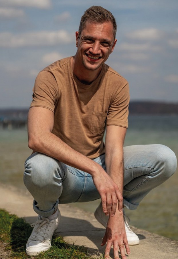 Dominik Hollop hockt lachend am Ufer, trägt ein braunes T-Shirt, Jeans und weiße Sneaker, im Hintergrund das Wasser.