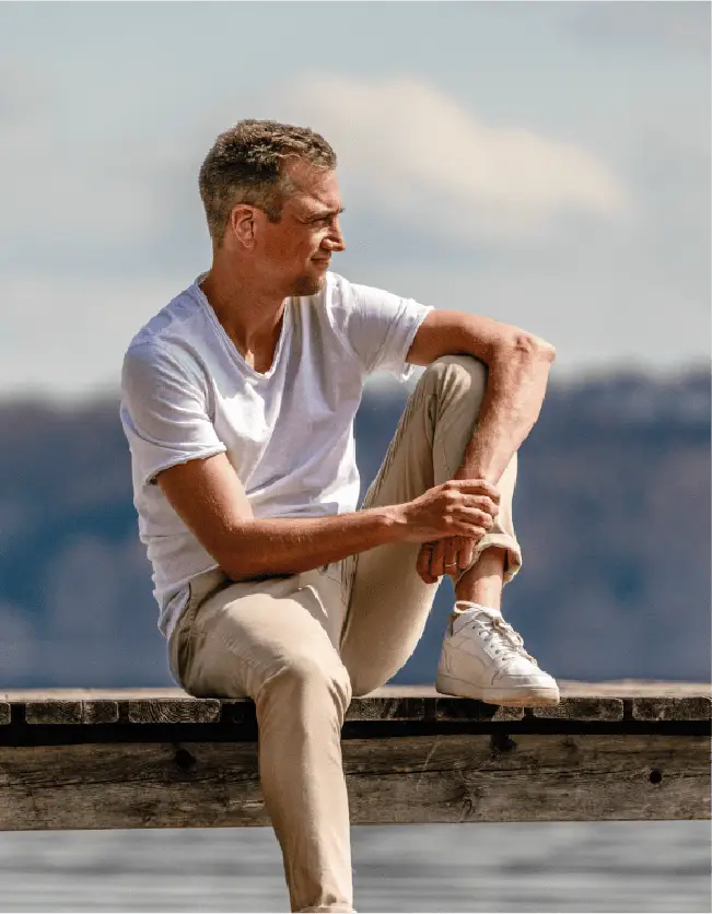 Dominik Hollop sitzt auf einem Holzsteg am Wasser, trägt eine Jeansjacke und helle Hose, und schaut lächelnd zur Seite.