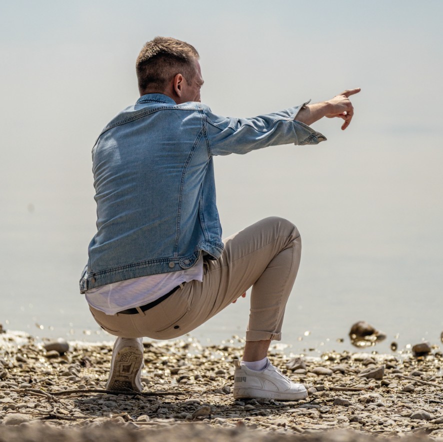 Dominik Hollop hockt am Seeufer, zeigt mit dem Finger nach vorne und trägt eine Jeansjacke mit heller Hose.