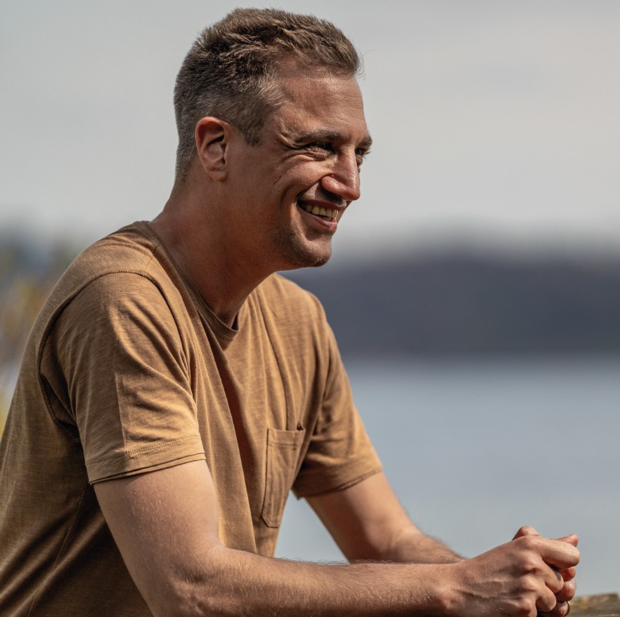 Dominik Hollop sitzt draußen an einem Tisch, lacht und schaut zur Seite, im Hintergrund See und Natur.