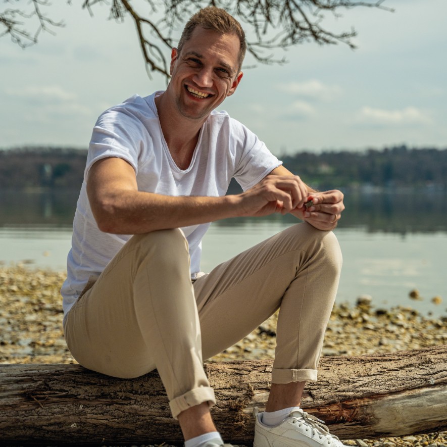 Dominik Hollop sitzt lächelnd auf einem Baumstamm am Seeufer, trägt ein weißes T-Shirt und helle Hosen