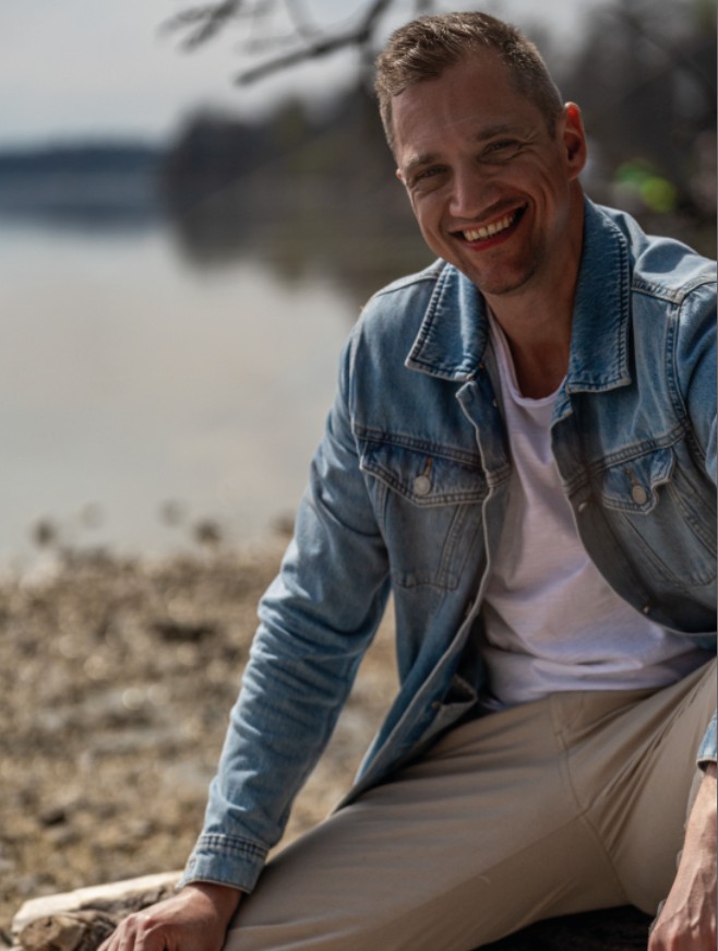 Dominik Hollop sitzt auf einem Steg, lächelt direkt in die Kamera, hinter ihm liegt der See im Sonnenschein.